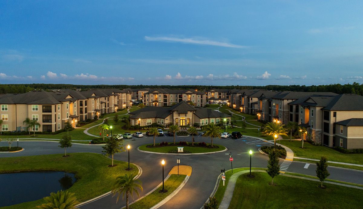 Angled view of the community showing the clubhouse surrounded by apartment buildings, and the street entrances with landscaping and lights