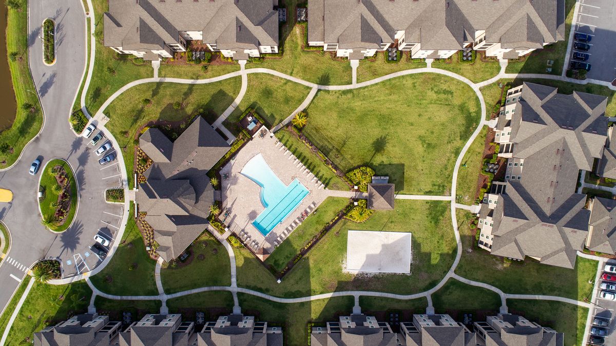 High up overhead view of the community showing apartment buildings that surround the clubhouse, pool courtyard, and sand volleyball court