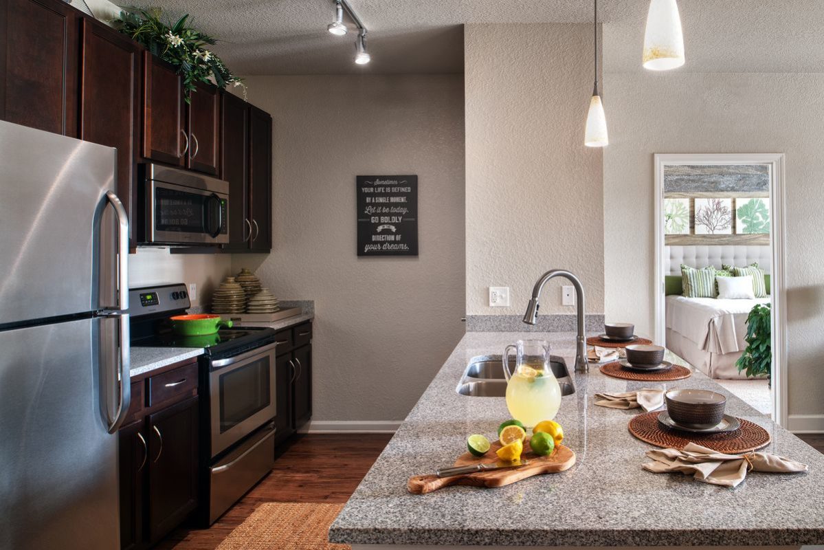 Apartment kitchen with wood flooring, track and pendant lighting, granite peninsula countertop with dual basin undermounted sink facing the living area, and stainless steel appliances